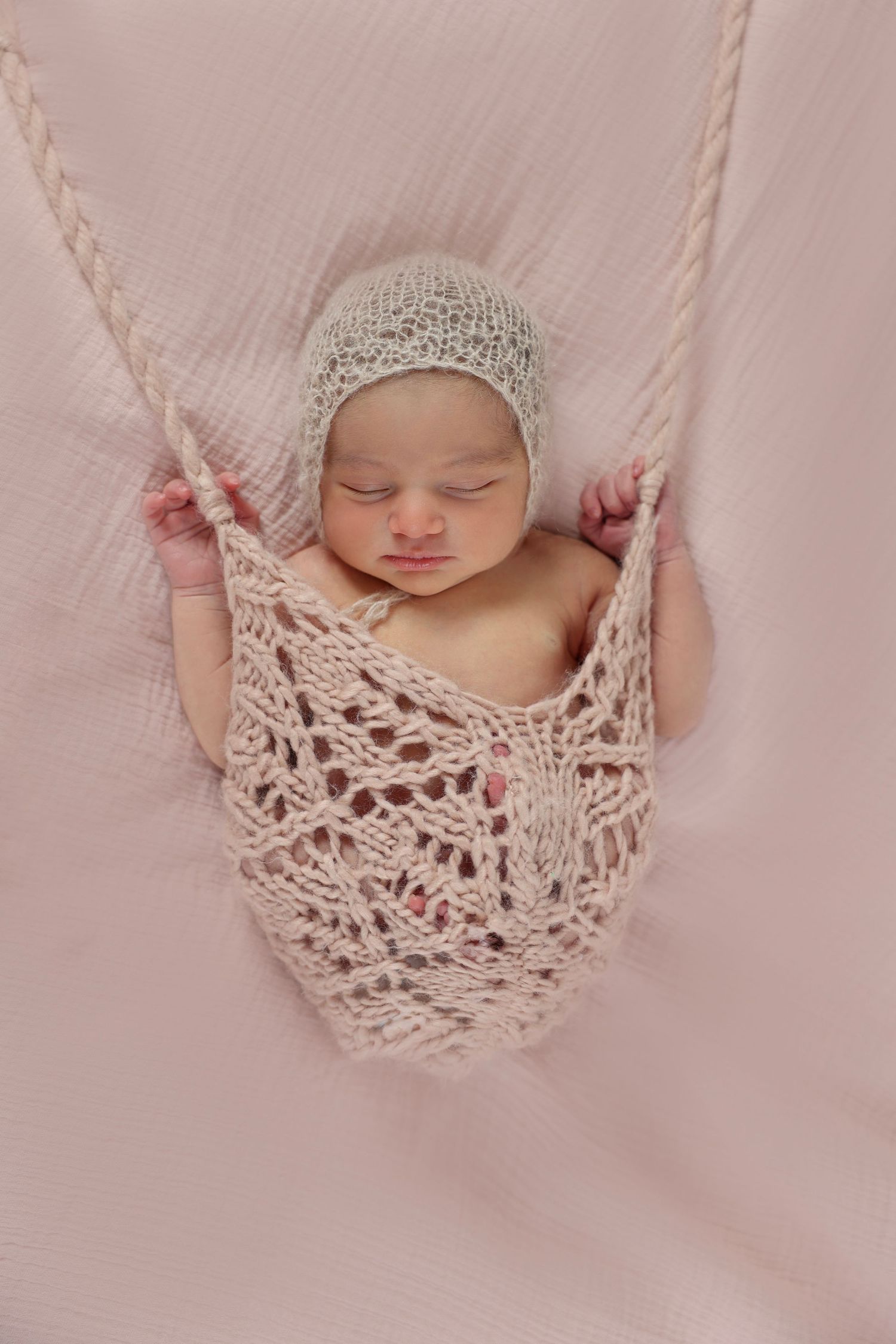 Sleeping newborn in a delicate, hand-knitted swing, wearing a matching hat on a soft pink background.