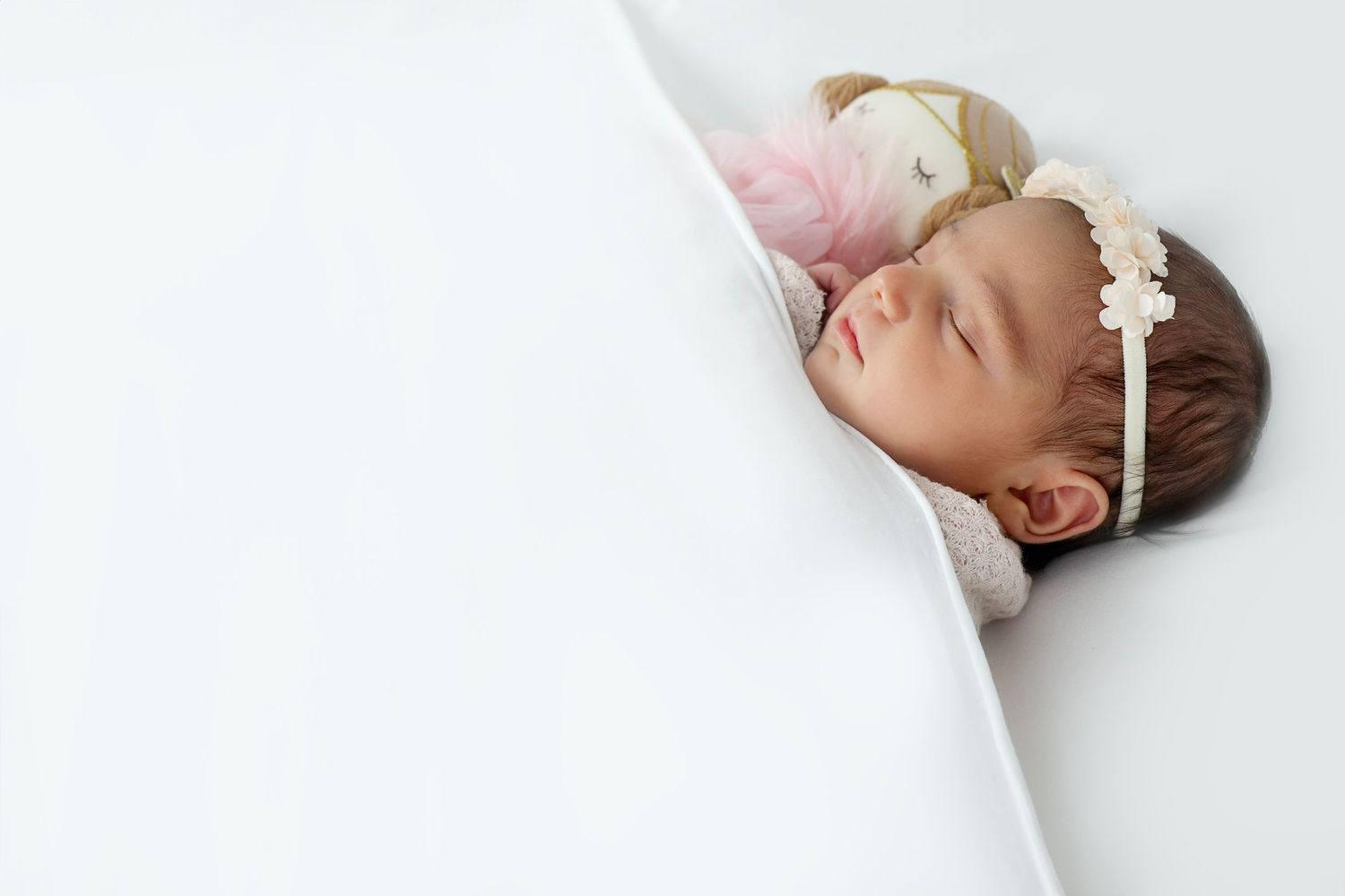 Peaceful newborn with a soft pink floral headband lying near a plush doll.