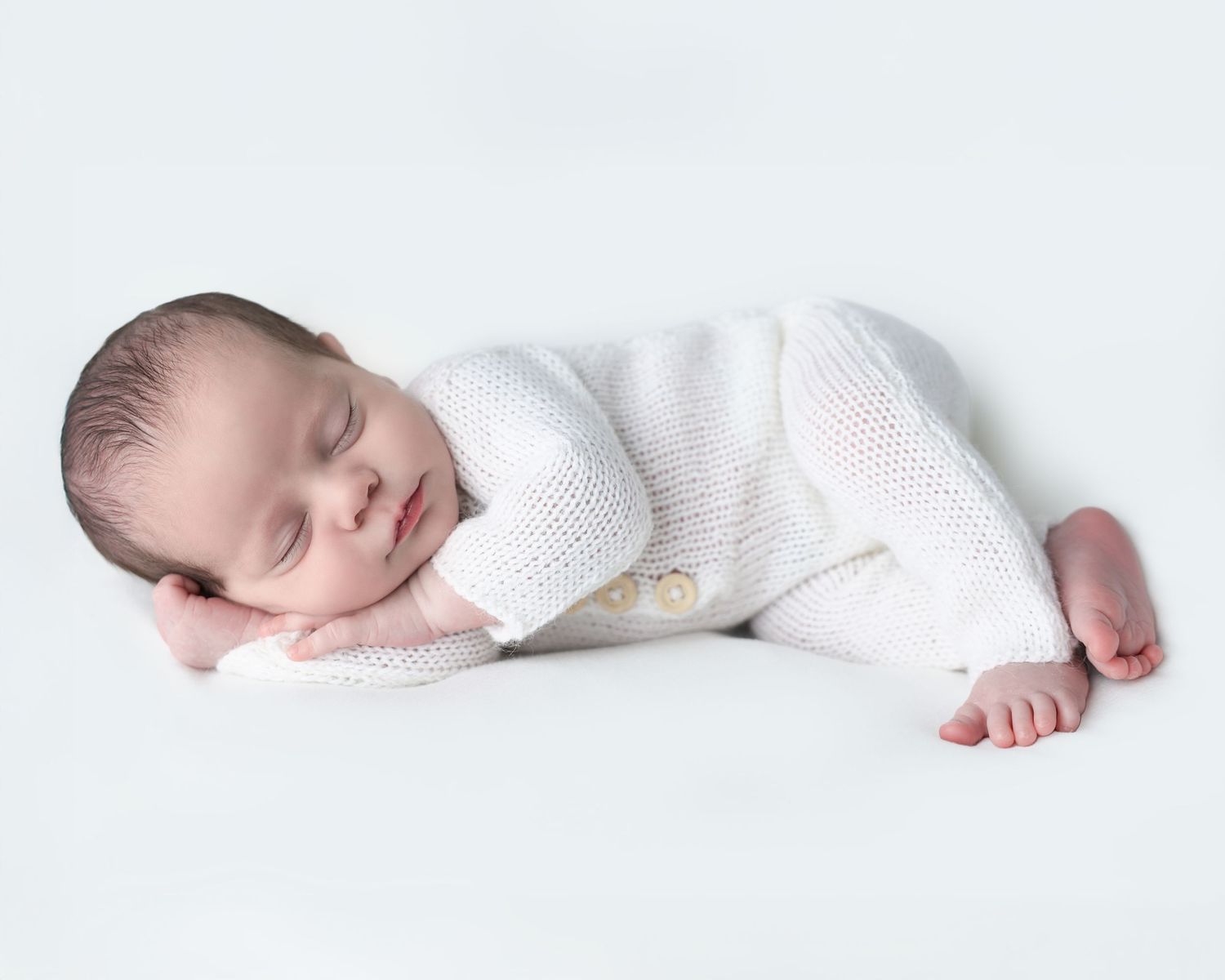 Minimalist newborn studio portrait of a sleeping baby wearing a white knit romper on a seamless white background