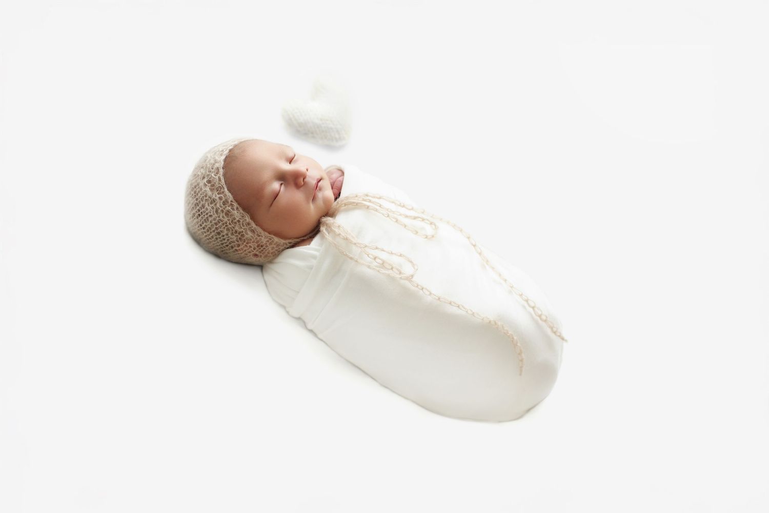 A peaceful newborn baby sleeping, wrapped in a white swaddle and wearing a delicate knit bonnet, lying on a soft white background