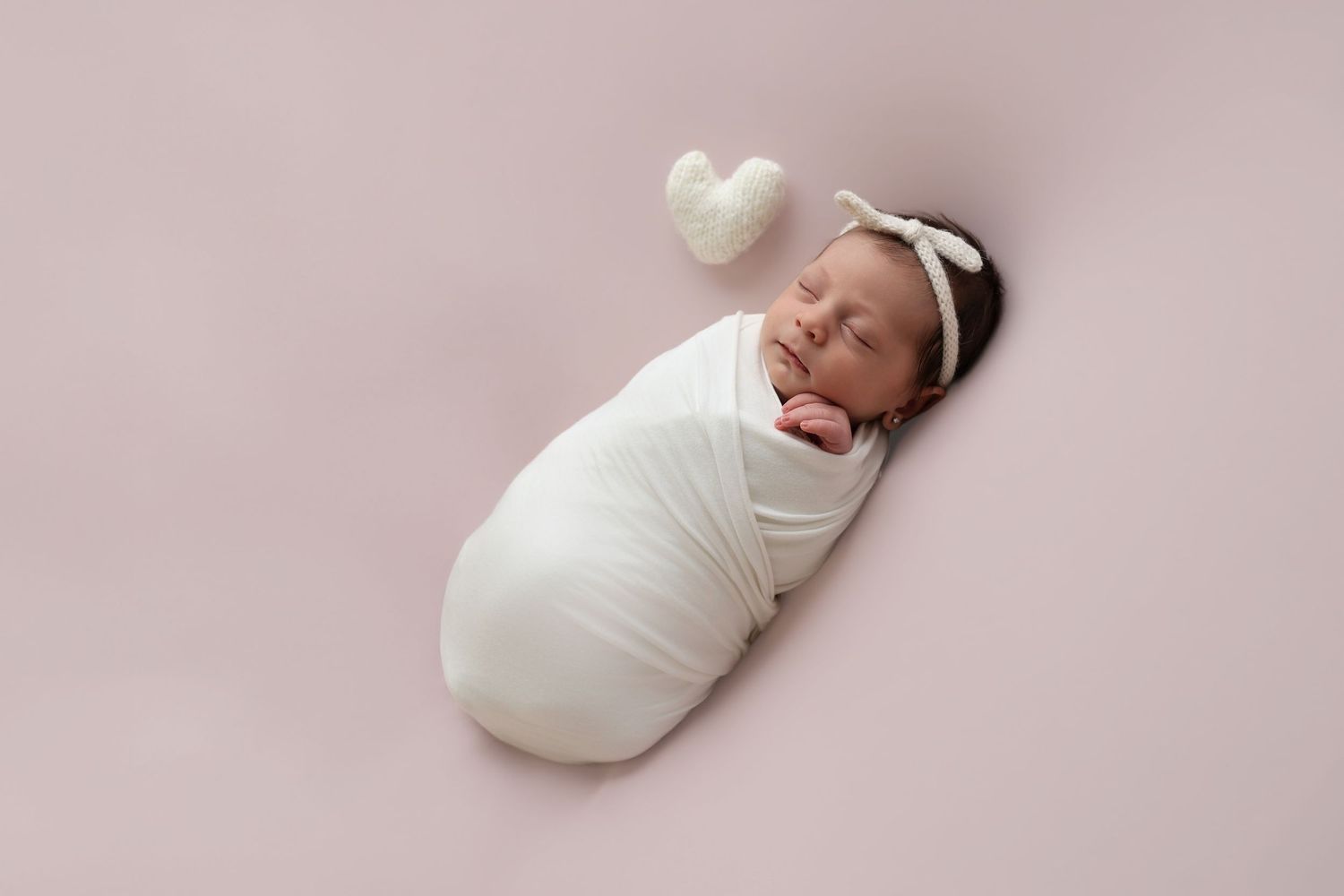 A peaceful newborn baby sleeping, wrapped in a white swaddle and wearing a knit bow, lying on a soft pink background