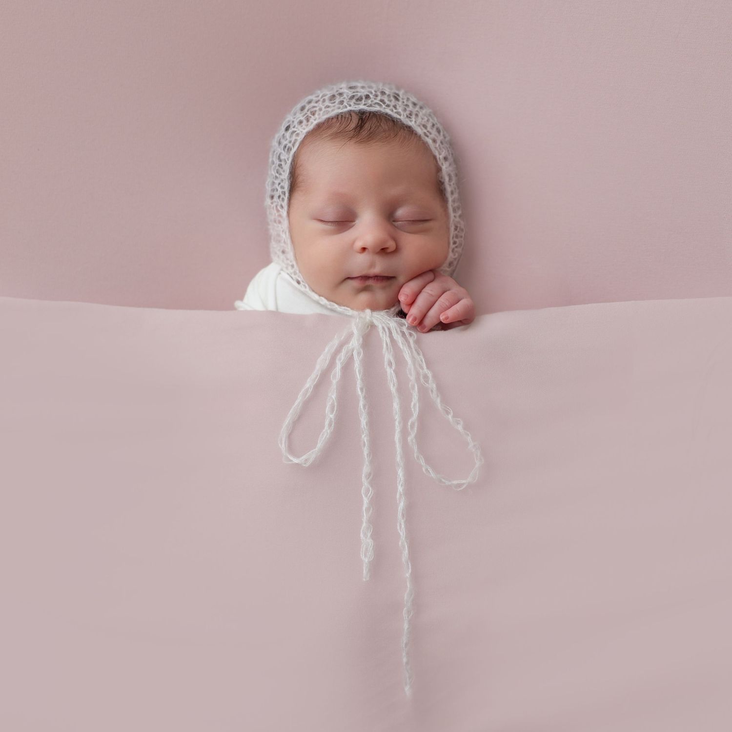 close up of newborn girl on a pink background wearing a lace bonnet with the bow tied over the pink blanket and her little hand peaking out