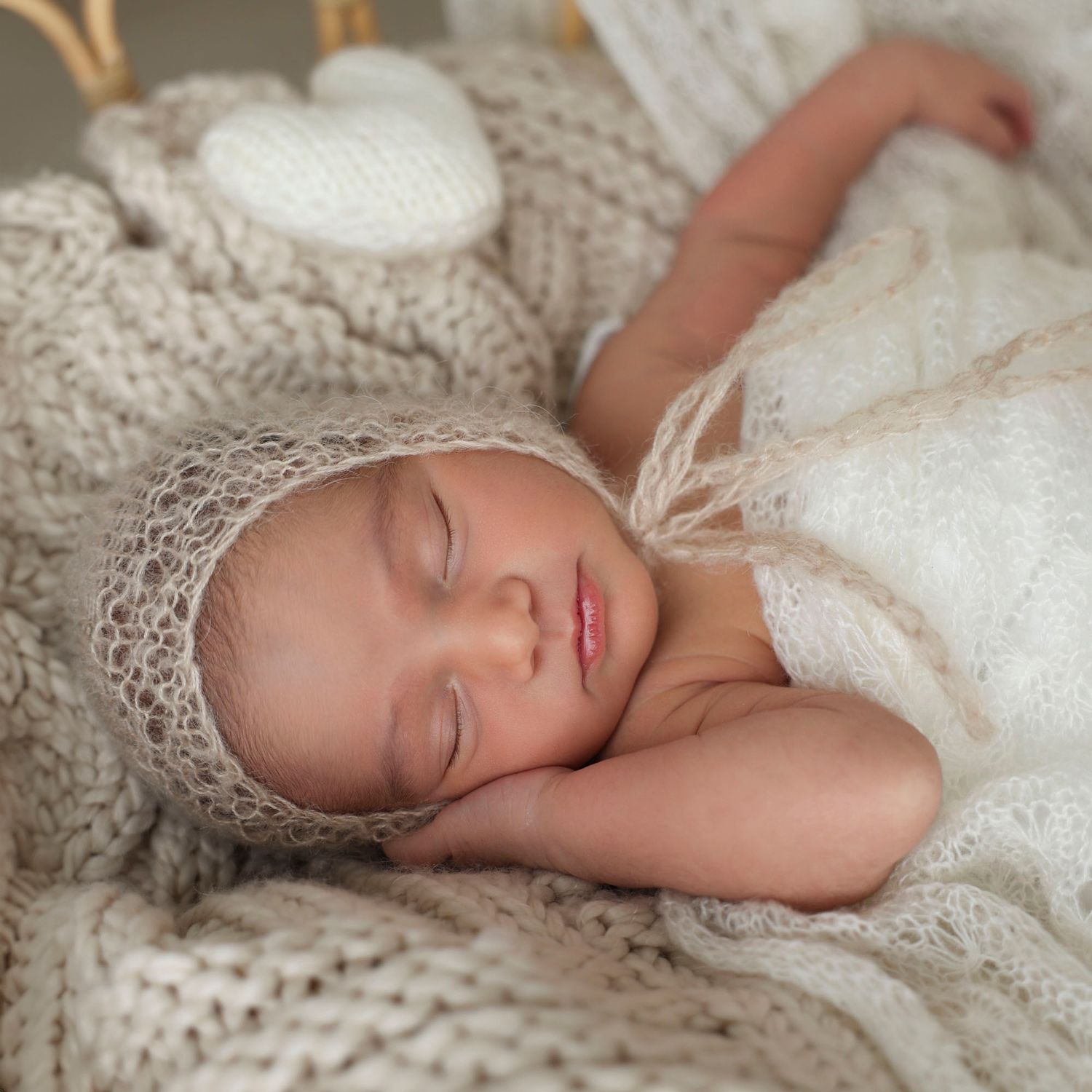 Sleeping newborn in a cozy, knitted bonnet and blanket, resting peacefully with a hand by the face against a neutral background.