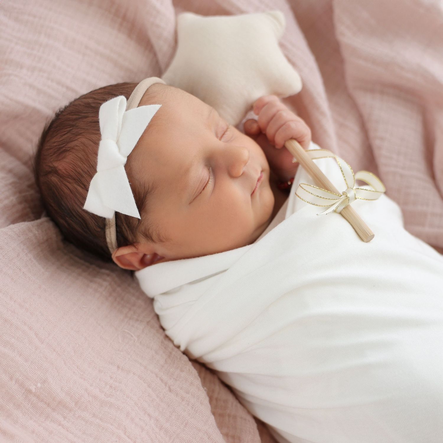 Sleeping newborn with a white bow is swaddled in a soft white wrap, her tiny hands holding a plush star