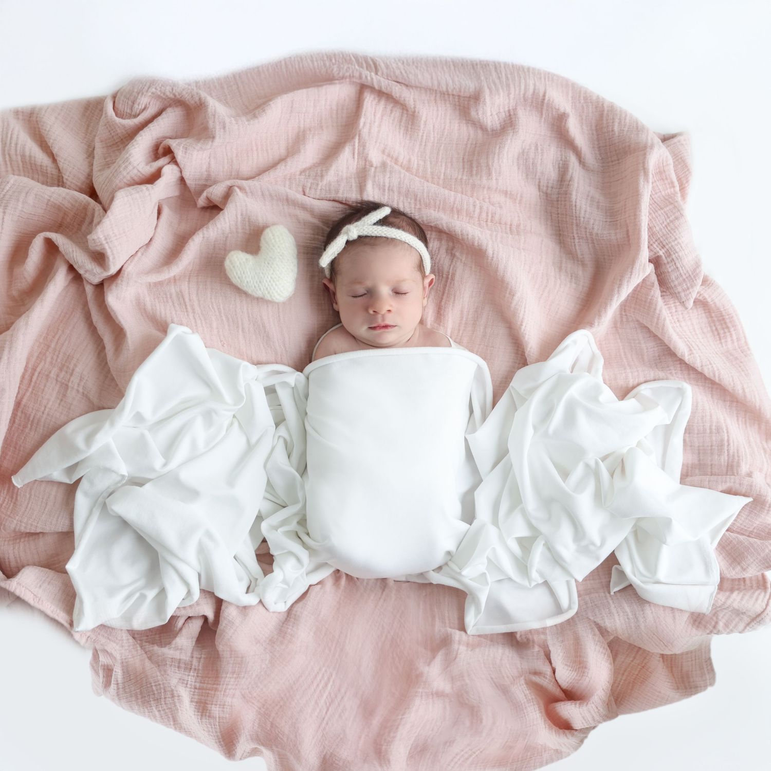 Newborn baby peacefully asleep on a soft pink blanket, wrapped in white fabric with a knitted heart nearby.