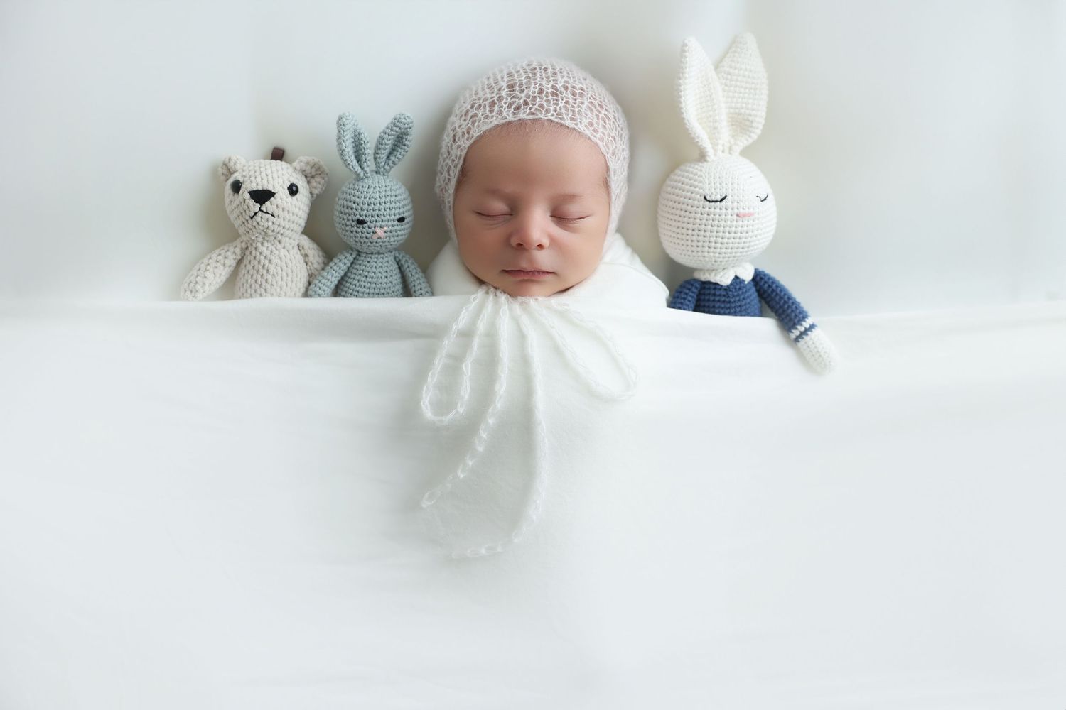 studio image of a newborn lying near soft knot teddy bears wearing a beautiful bonnet