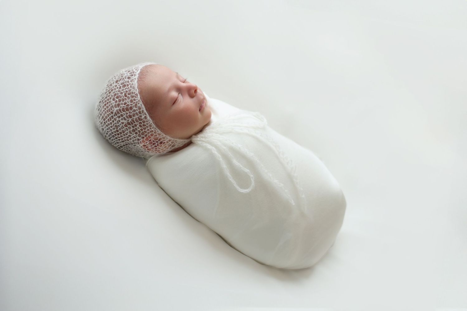 A peaceful newborn baby sleeping, wrapped snugly in a white swaddle and wearing a bonnet, lying on a soft white background