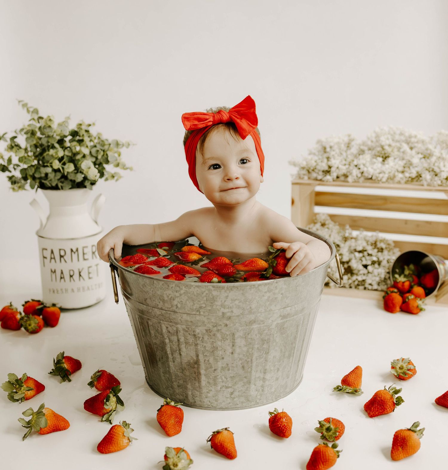 A baby wearing a red bow sits in a metal tub surrounded by fresh strawberries for a styled photoshoot.