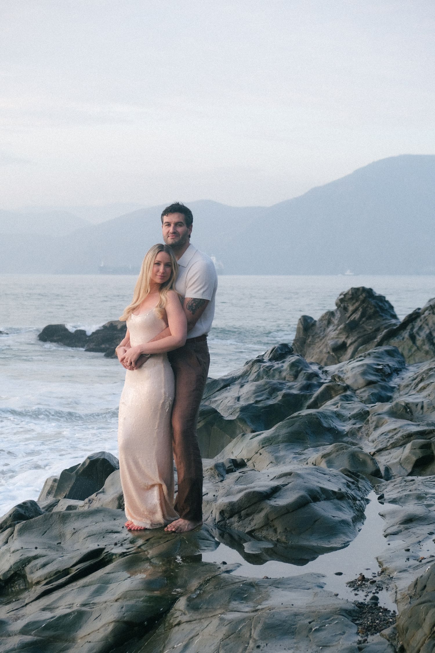 A couple embraces on a beach with the Golden Gate Bridge visible in the background.