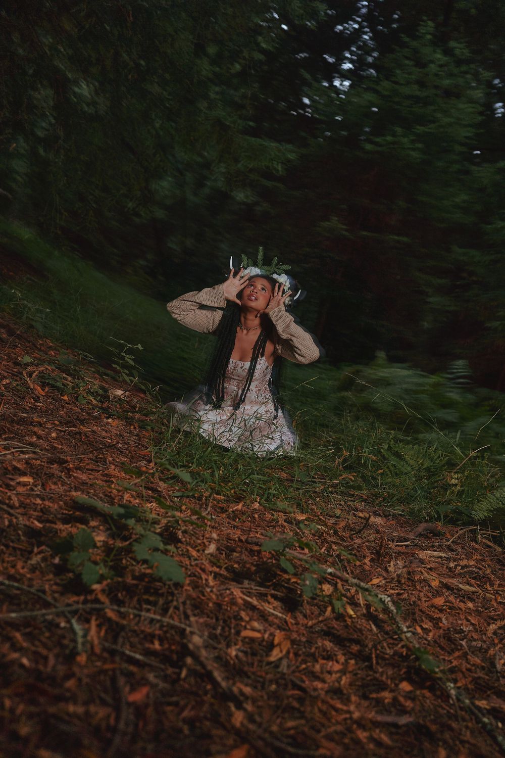 Woman in floral dress with antler crown sits on forest floor surrounded by dark trees for a fairy  photoshoot in Hayward, CA.