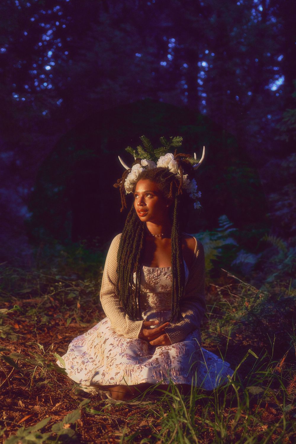 Woman in floral dress with antler crown kneels in dark forest illuminated by warm light for a fairy photoshoot in Hayward, CA.