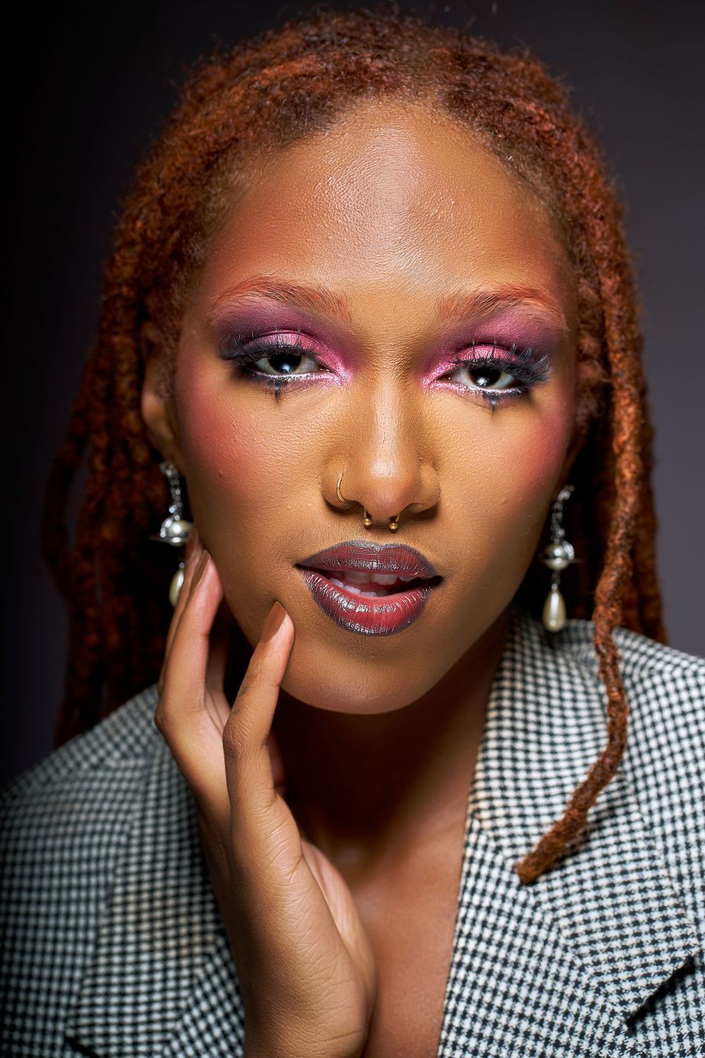 A Black woman with auburn braids wearing pearl earrings and bold pink eye makeup poses in a houndstooth blazer for a beauty shot in Oakland.