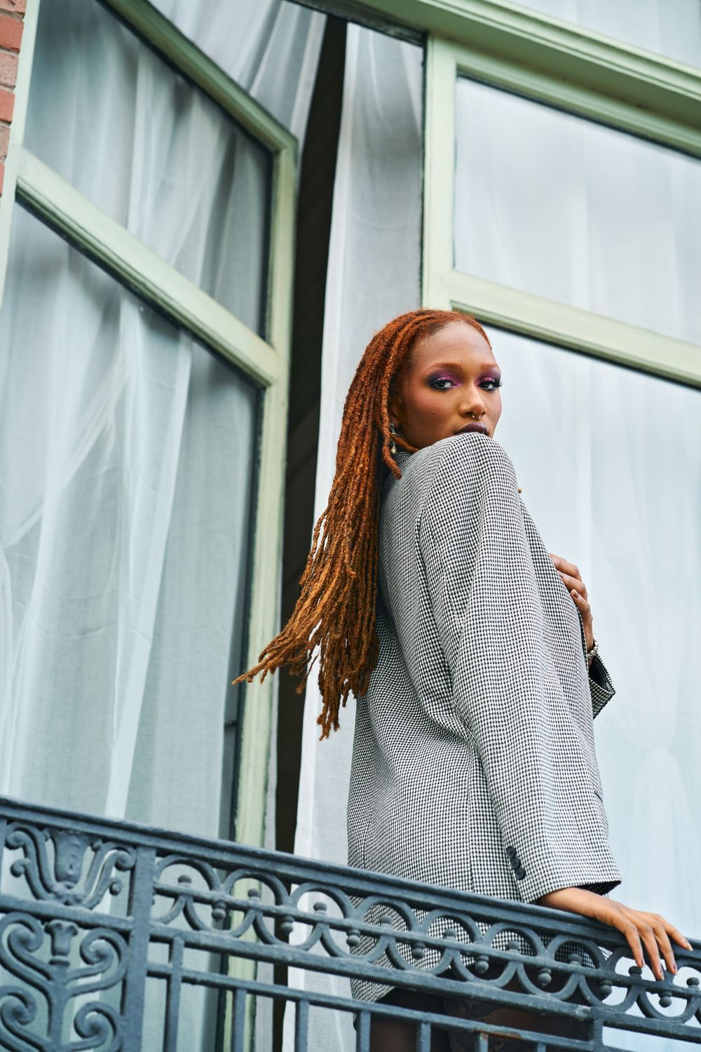 A Black woman with long auburn braids wearing a grey blazer stands on an ornate balcony during a photoshoot in Oakland, CA.