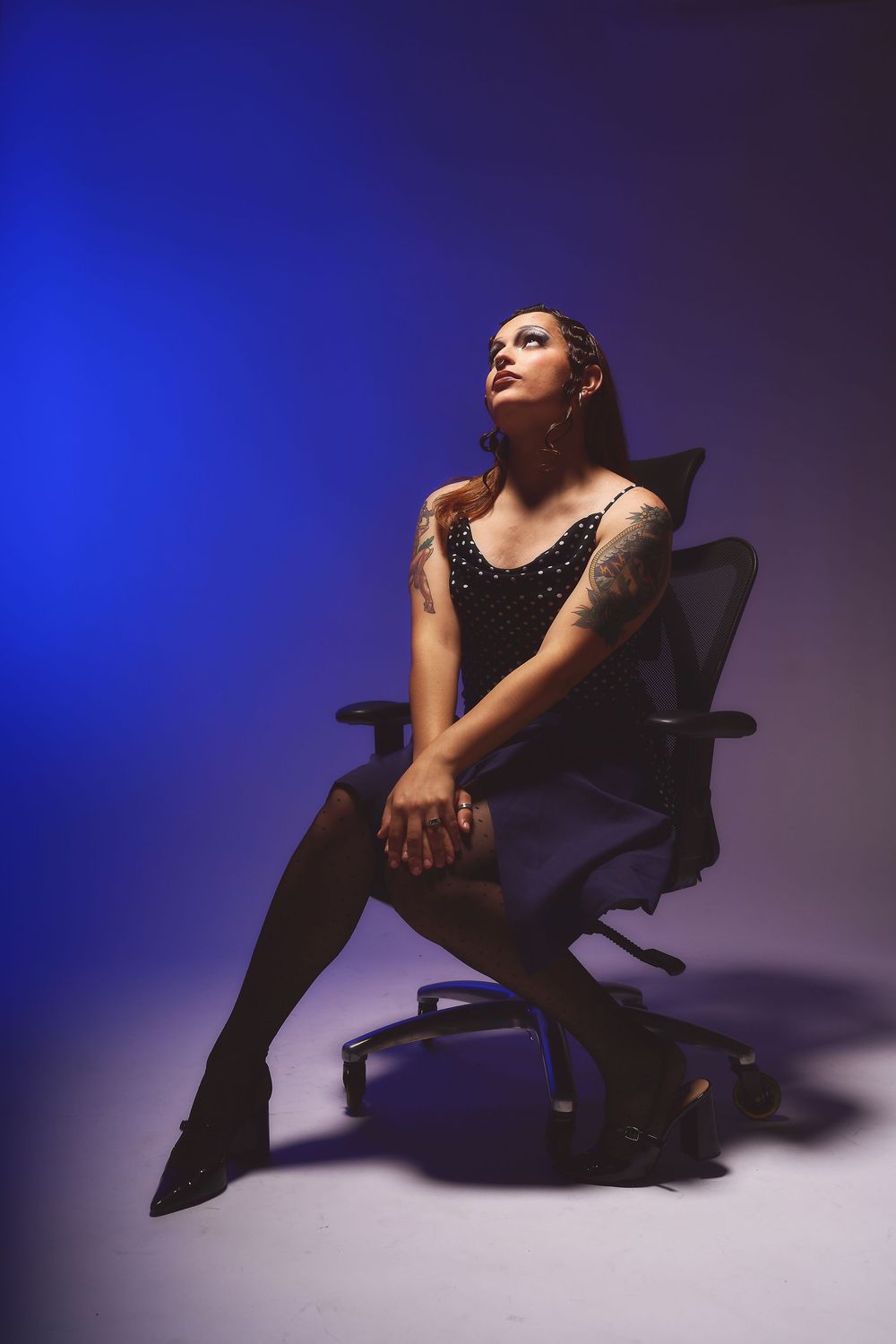 Tattooed woman in black dress and thigh-highs poses in office chair against blue-purple backdrop for an editorial photoshoot in Oakland.