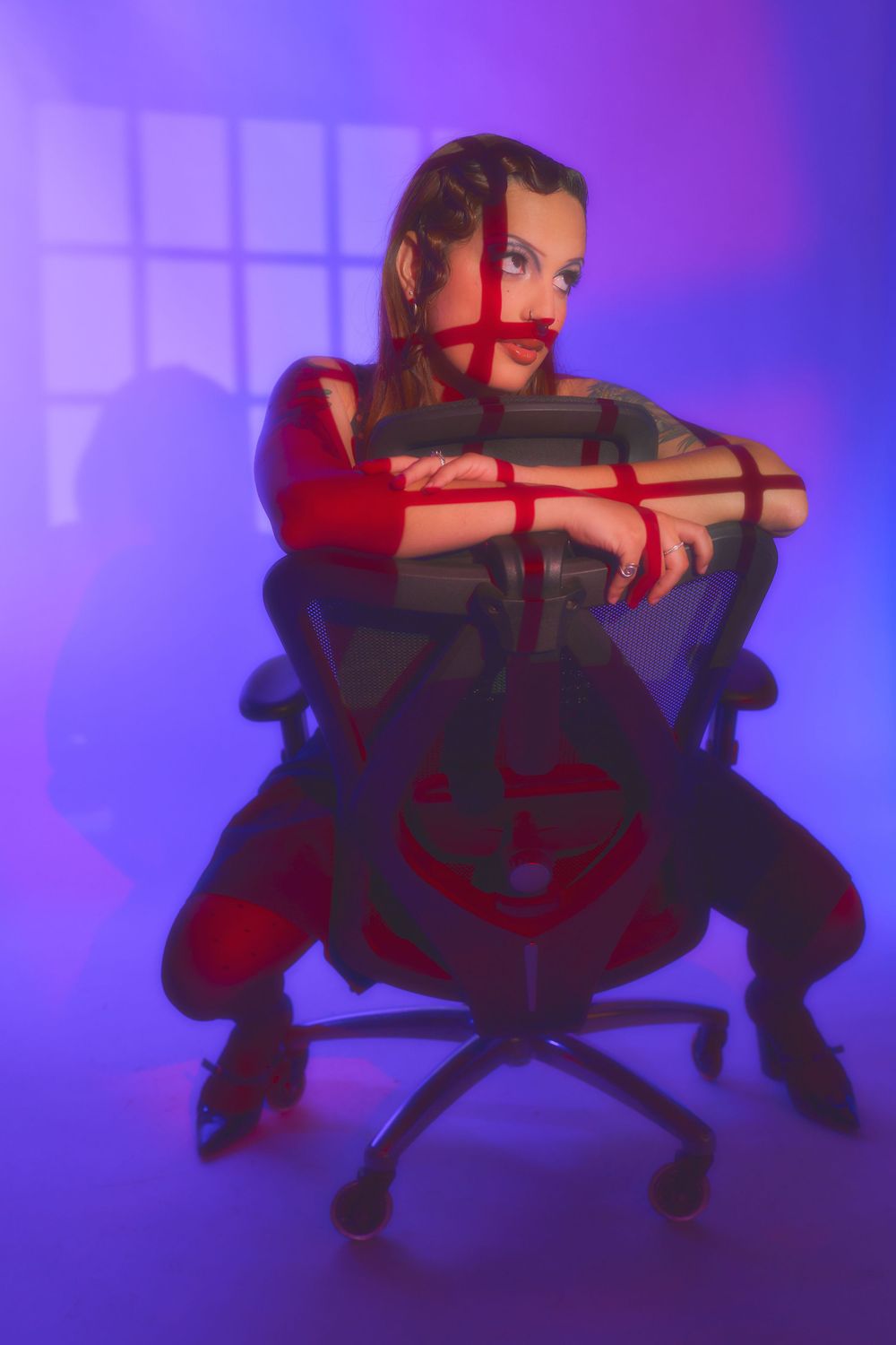 Woman bound in red rope sits in office chair under dramatic purple and red studio lighting for an editorial photoshoot.