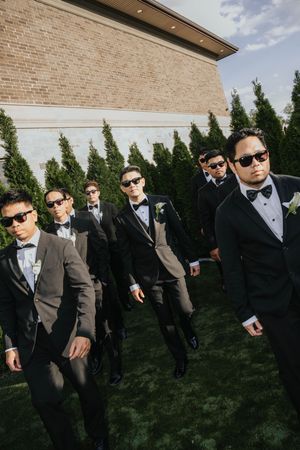 Groomsmen in black tuxedos and sunglasses pose together on grass in front of brick building wall.