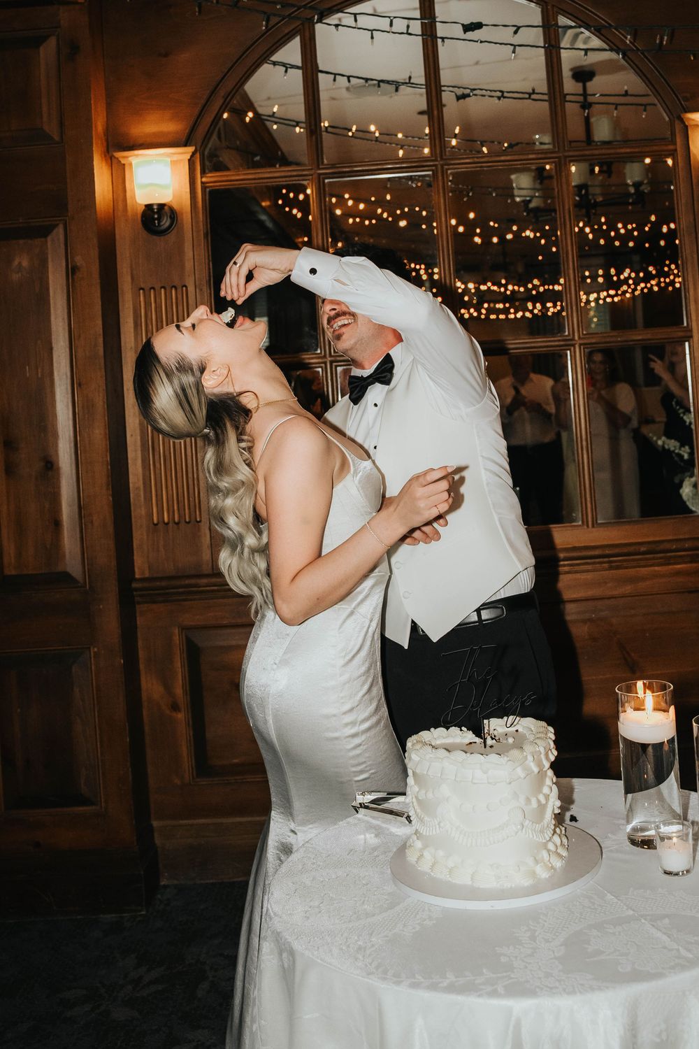 Newlywed couple shares a playful moment while cutting their white wedding cake at an indoor reception with twinkling lights.