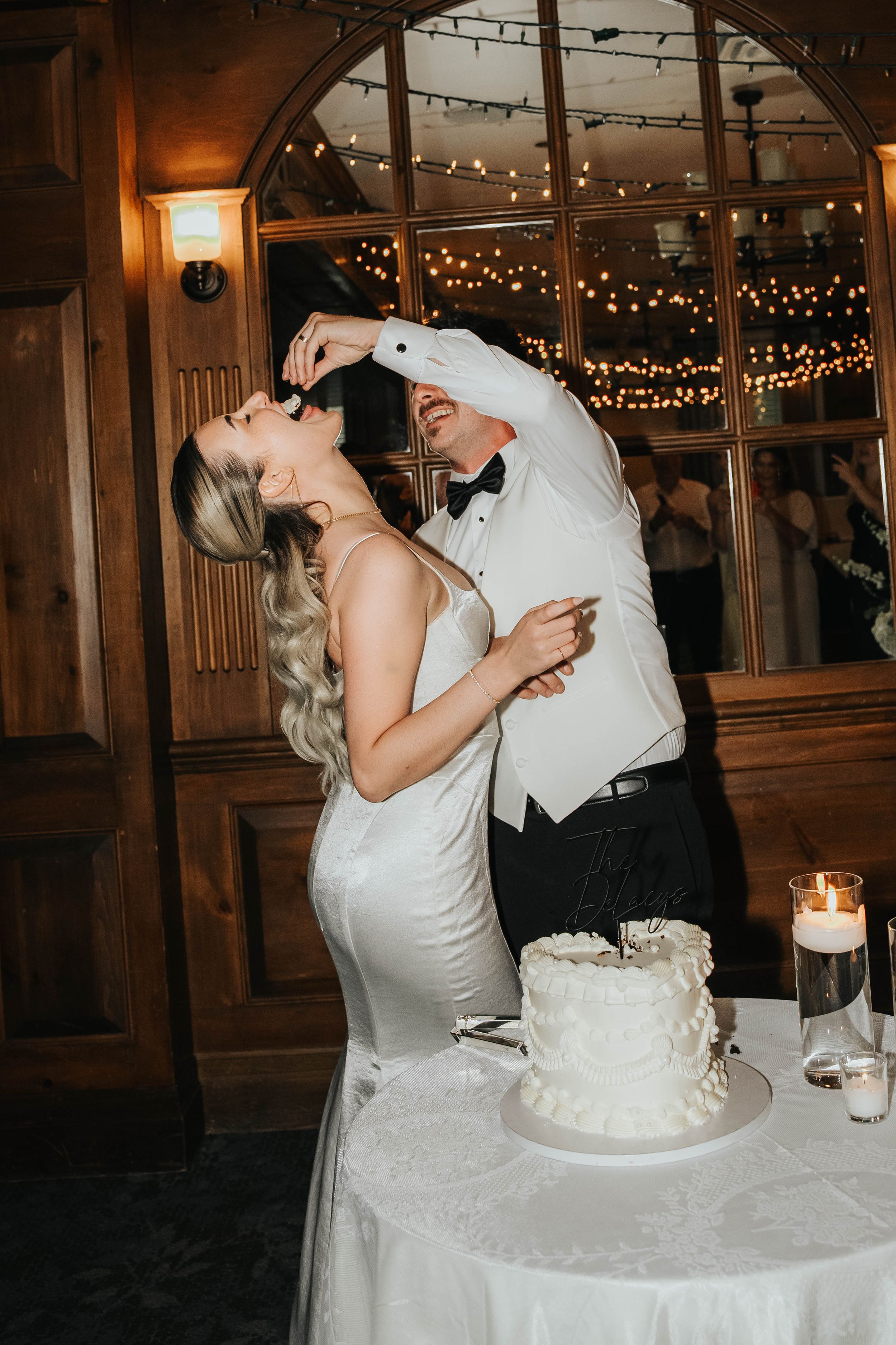 Newlywed couple shares a playful moment while cutting their white wedding cake at an indoor reception with twinkling lights.
