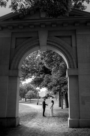 A black and white photo of two people standing beneath a stone archway on a tree-lined path.