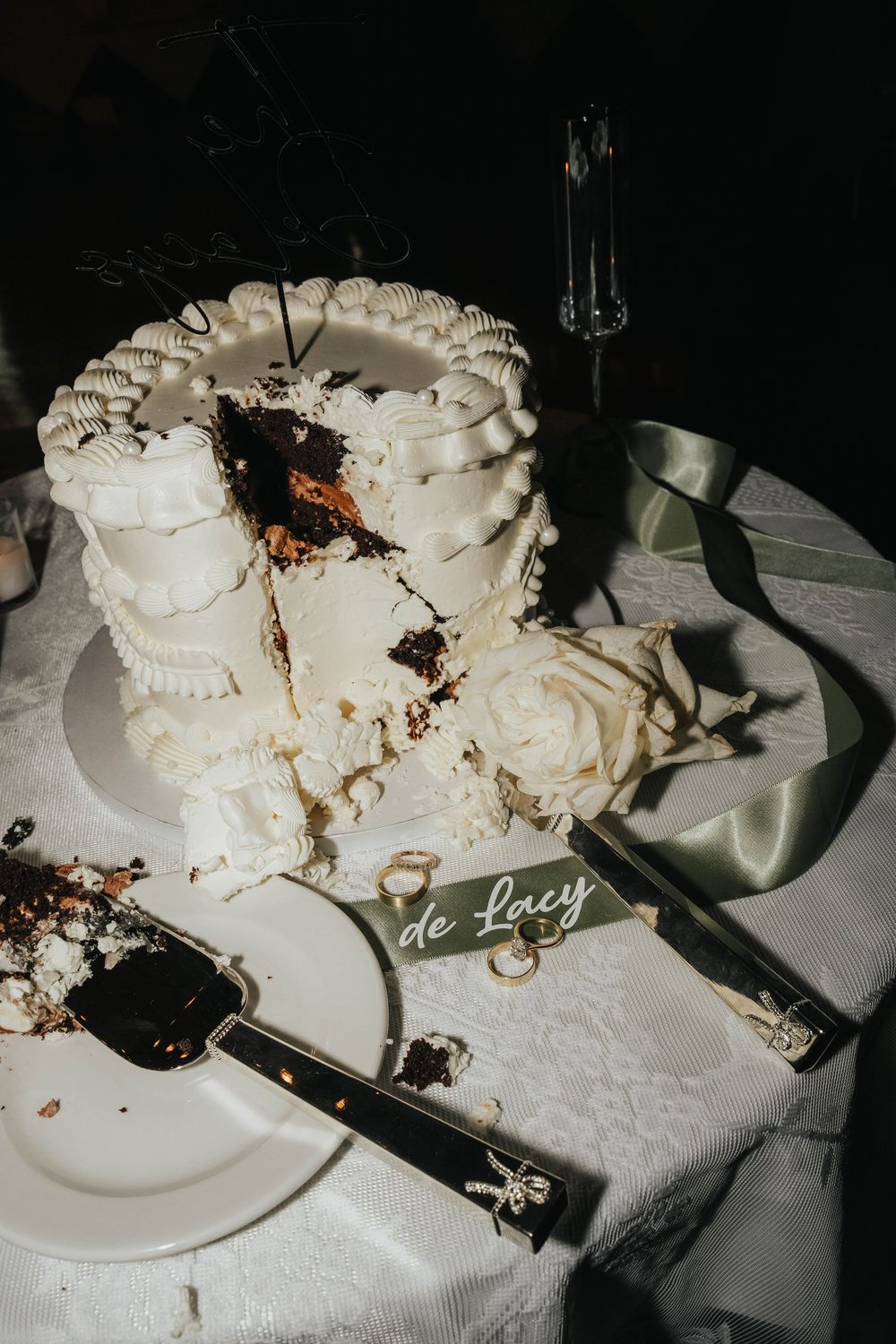 A sliced white wedding cake with ruffle details sits on a table with serving utensils and a dark background.