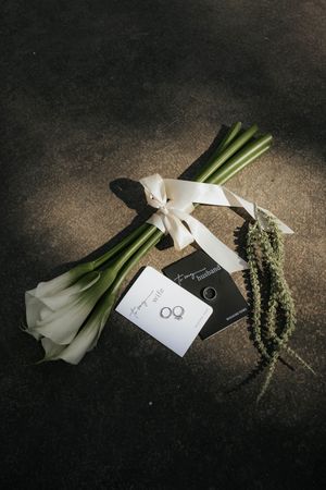 White calla lilies with green stems and wedding details arranged artistically on a dark background.