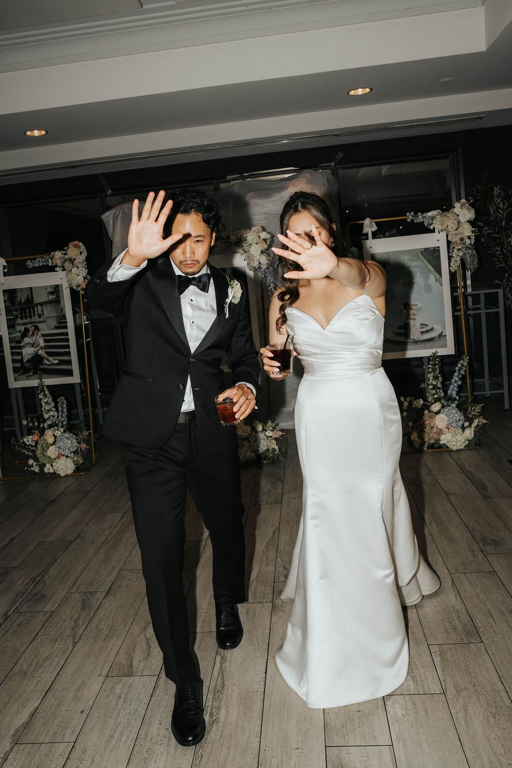 A couple in formal wedding attire dances together on a light wooden dance floor with floral decorations in the background.