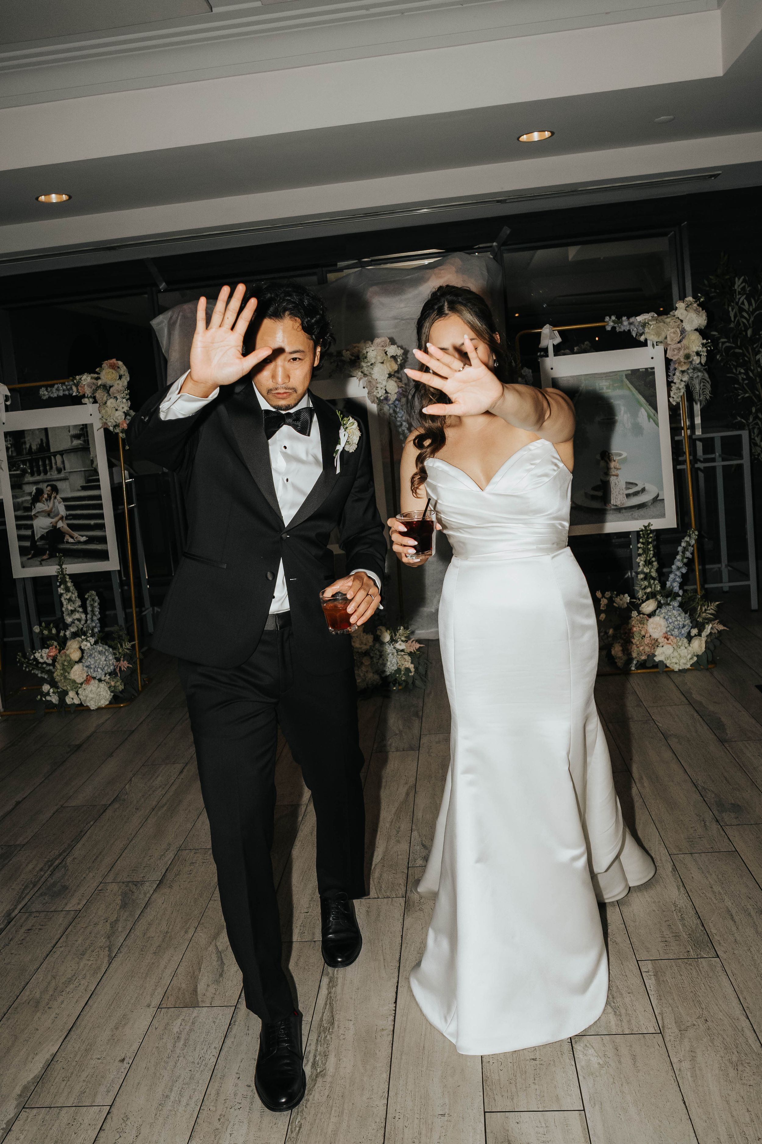 A couple in formal wedding attire dances together on a light wooden dance floor with floral decorations in the background.
