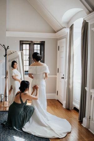 Wedding dress preparation scene in elegant white room with bride looking in full-length mirror.