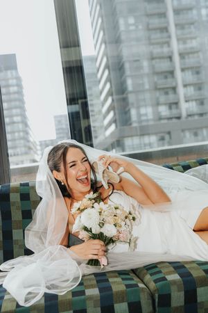 A bride in a white wedding dress lounges on a green striped couch with a bouquet of pink and white flowers.