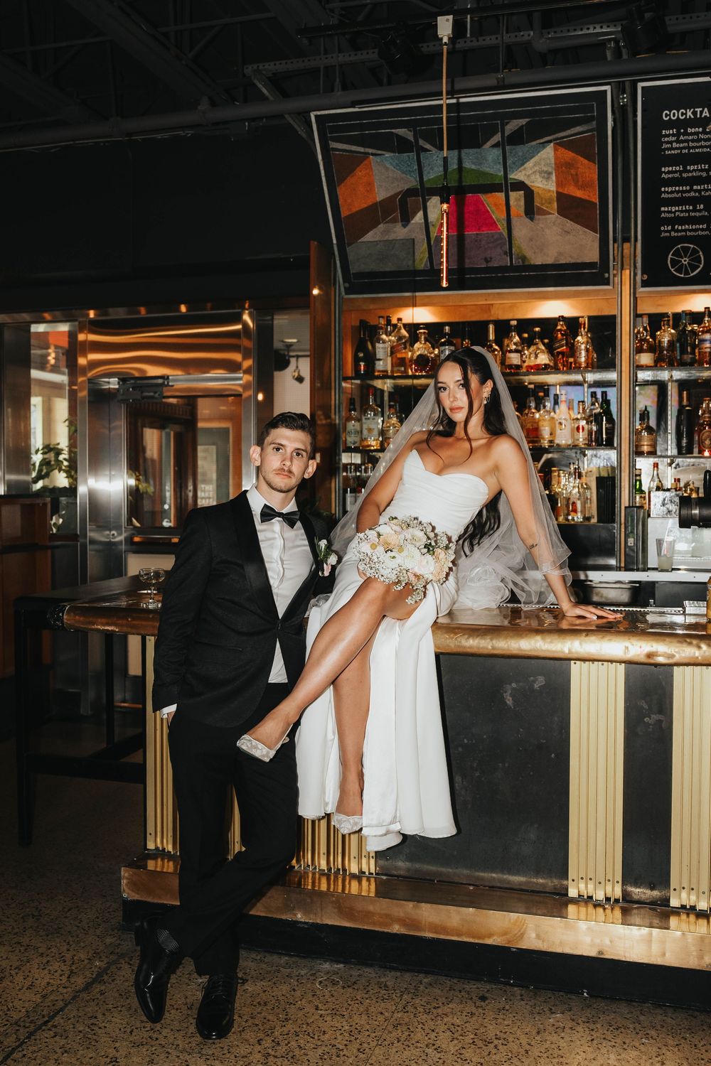 A couple in formal wedding attire poses playfully at an elegant bar with gold accents and warm lighting.