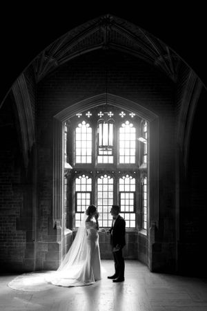 Silhouettes stand in front of a grand gothic window in a dramatic black and white photograph.