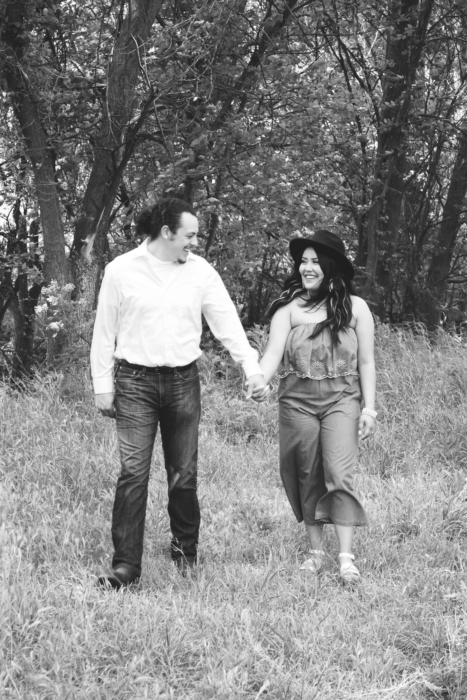 A romantic black and white photo of a couple walking hand in hand through a wooded area.