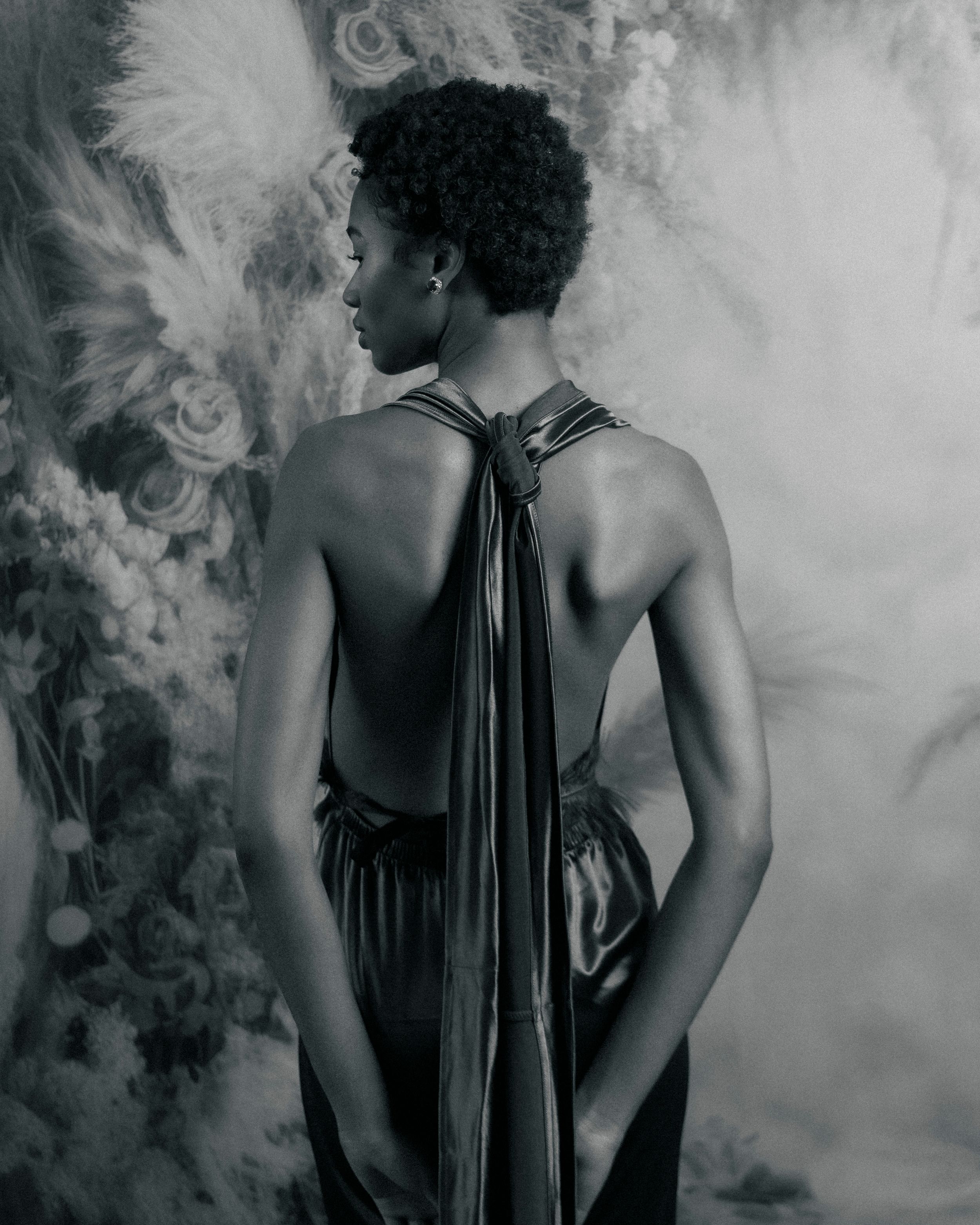 Profile view in black and white showing draped fabric and strong shoulder lines against ethereal backdrop.
