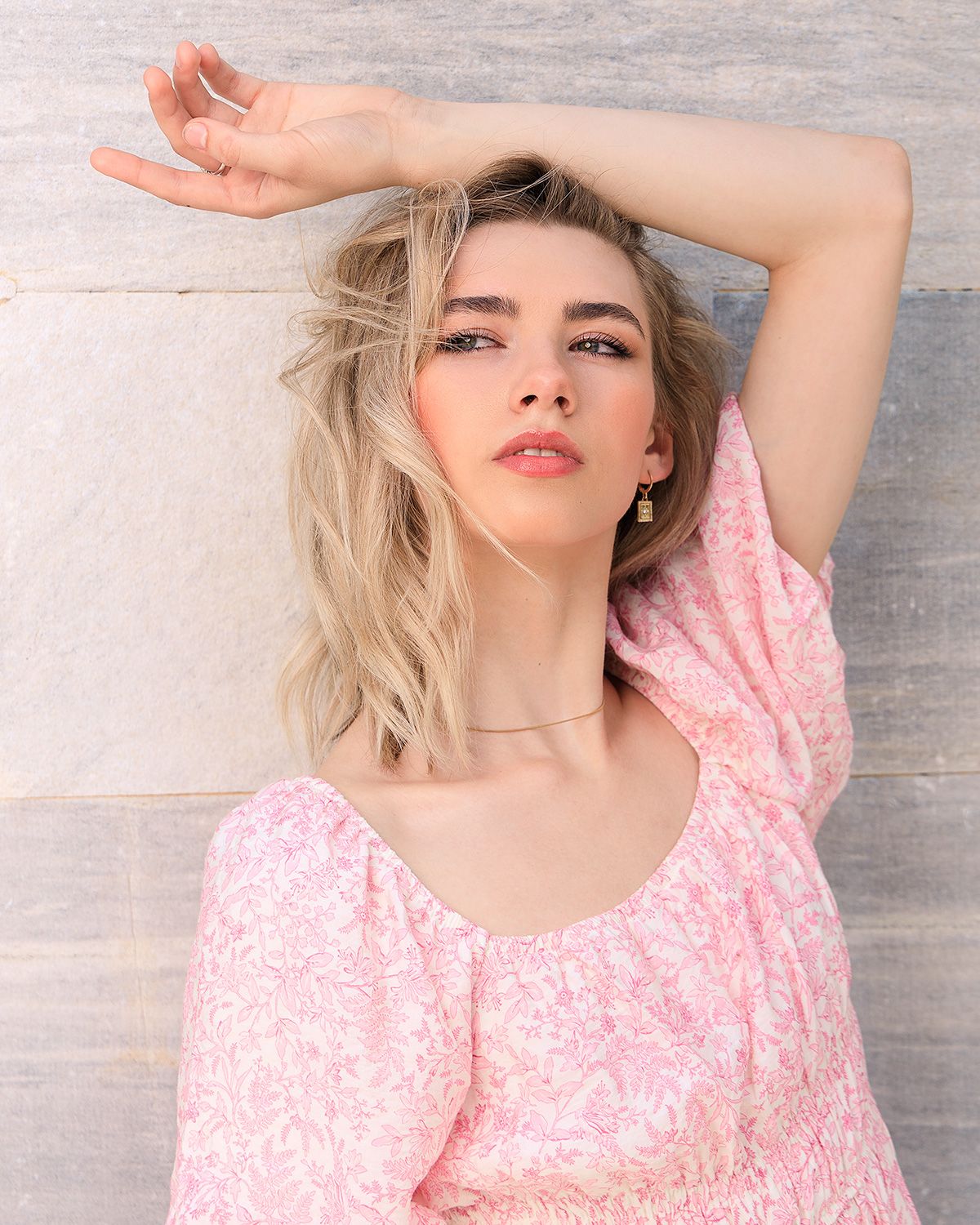 Fashion portrait in pink floral dress with arm raised against neutral wall.