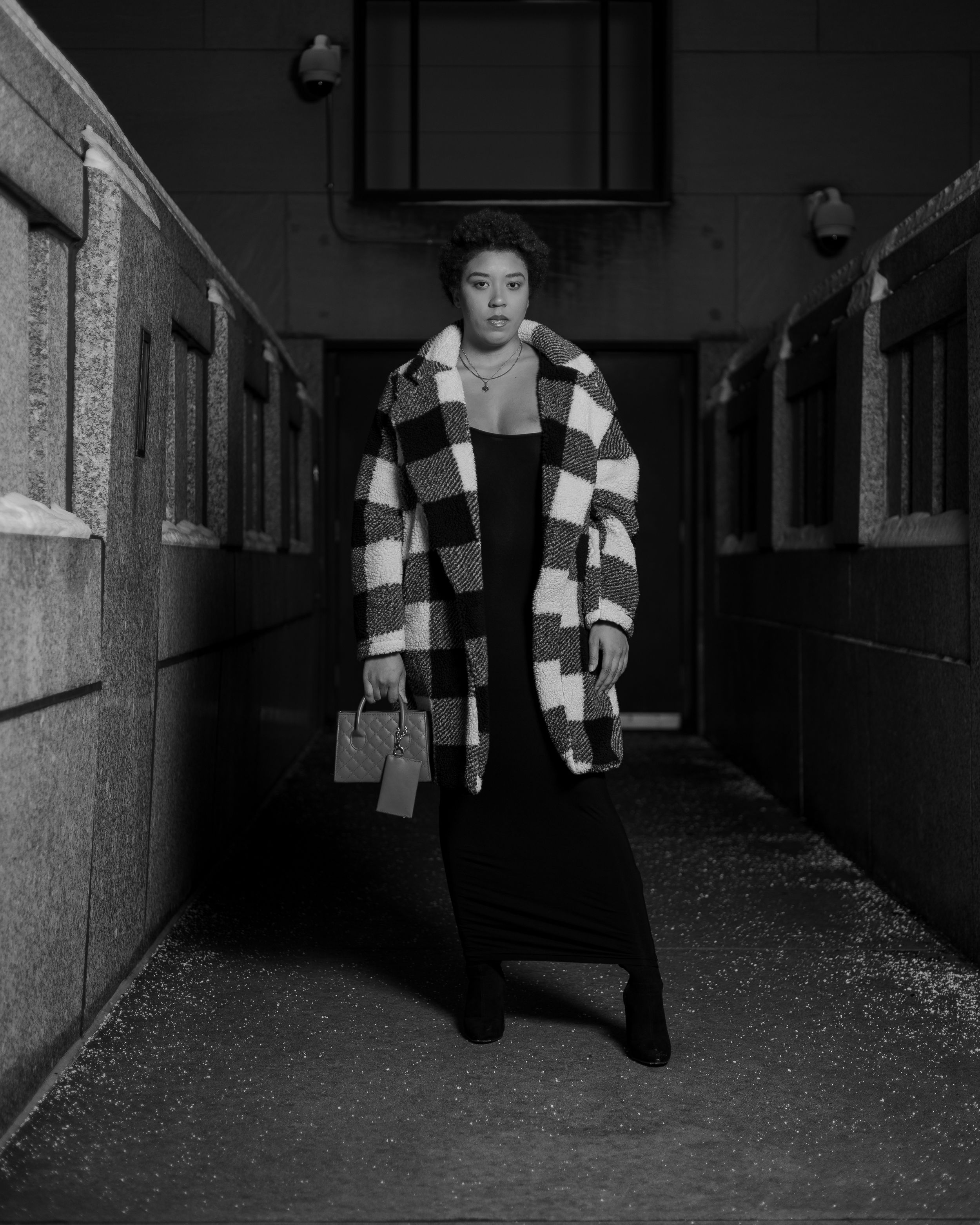 Black and white fashion photo in checkered coat standing in industrial hallway.