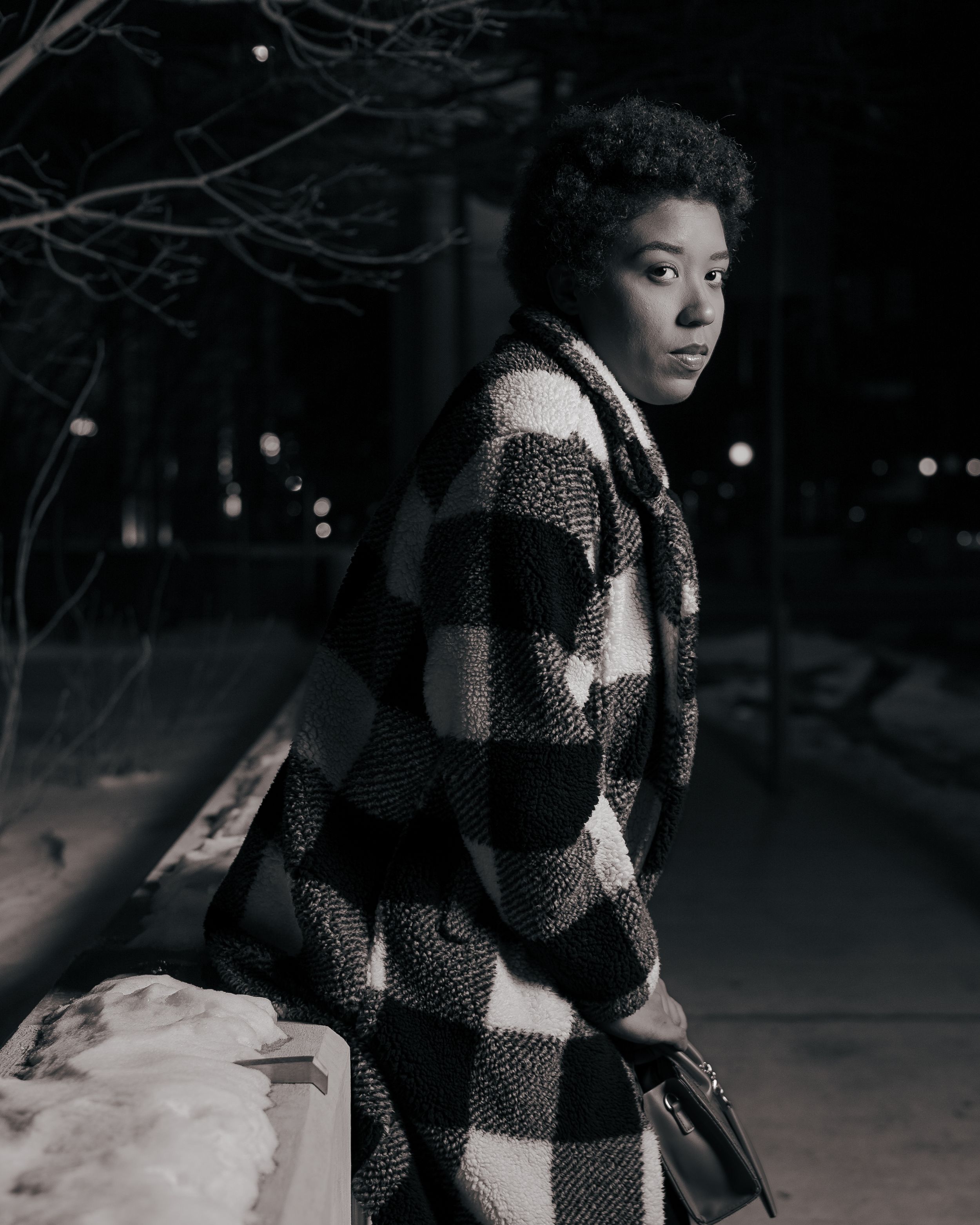 A moody black and white portrait showing a person in a checkered coat against a dark winter background.