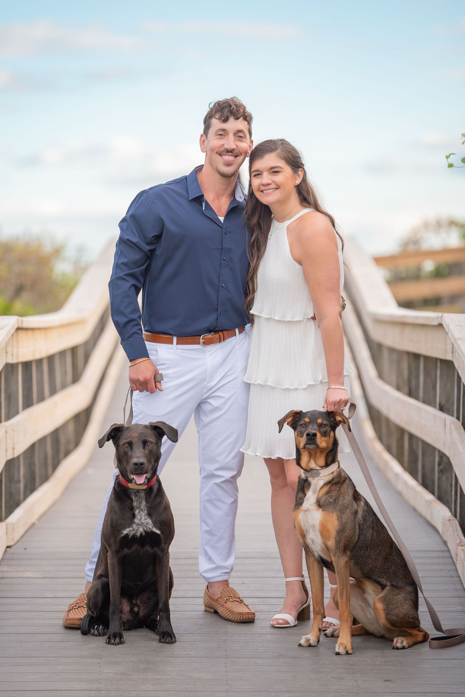 Couple posing with their three dogs on wooden boardwalk by the beach.