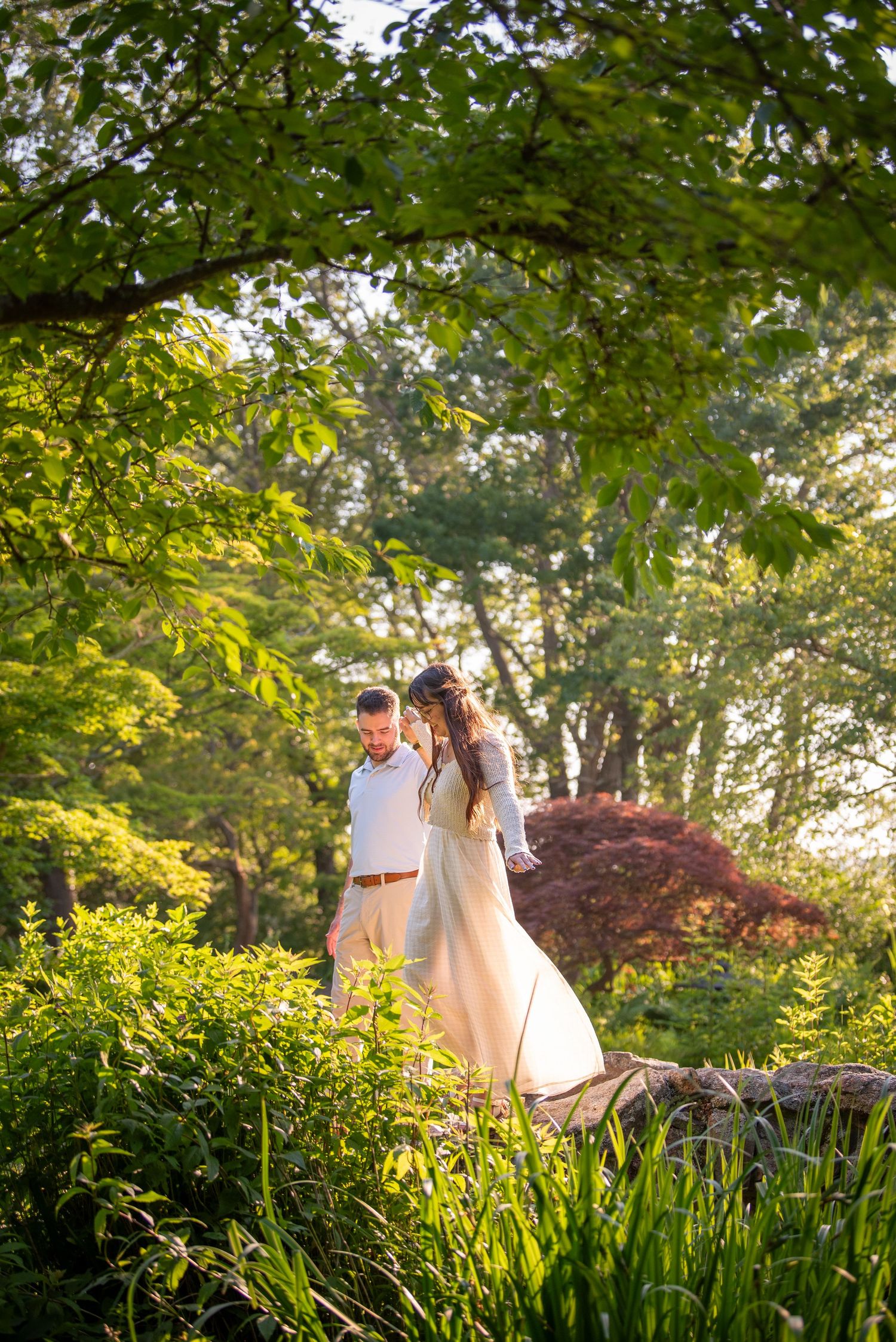 Couple in wedding attire walking through a lush garden path during golden hour with green foliage overhead.