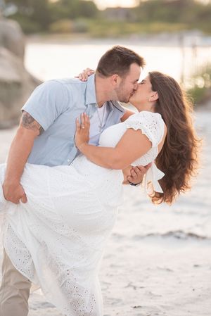 Romantic beach photoshoot with couple in white outfits embracing on sandy shore at sunset.
