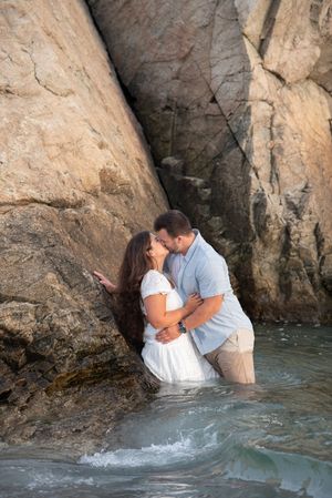 Couple sharing intimate moment while standing in ocean water against rocky coastal cliffs.
