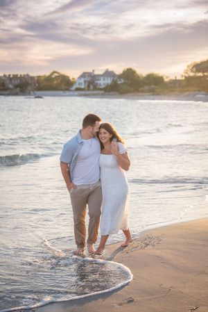 Couple walking along beach shoreline at sunset with coastal homes in background.