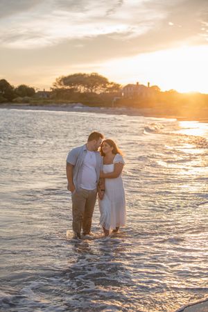 Couple standing in gentle ocean waves during beautiful golden sunset with coastal homes in background.
