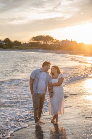 Couple walking along shoreline during sunset with waves lapping at their feet.