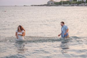 Playful couple wading in ocean water during coastal photoshoot at dusk.