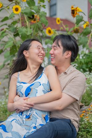 A romantic couple shares joyful laughter while sitting together among blooming sunflowers in a garden.