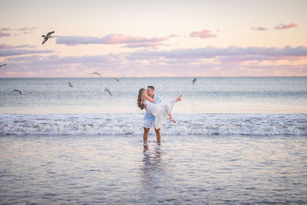 A romantic couple embraces while standing in shallow ocean water at sunset as seagulls fly overhead.