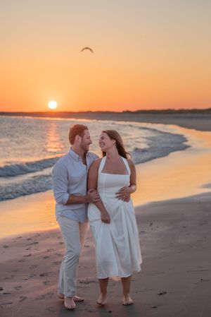 A couple in white outfits walks along a beach at sunset with waves and a seabird in the background.
