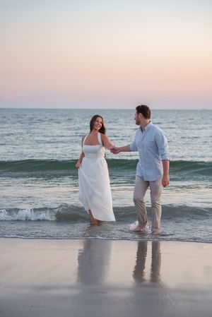 A couple walks together through shallow ocean waves during a sunset beach photoshoot.
