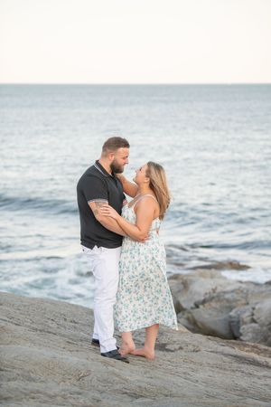 A romantic couple embraces on rocky coastal terrain at sunset with ocean waves in the background.