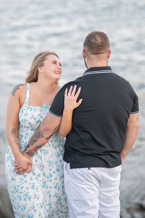 A couple shares a playful moment by the ocean, with the woman in a floral dress laughing while touching her partner's back.